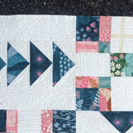 A close-up of a colorful Crosswind Mystery Quilt reveals geometric patterns, featuring blue triangles and patchwork squares in pink, green, and yellow on a white background, lying on a dark textured surface.