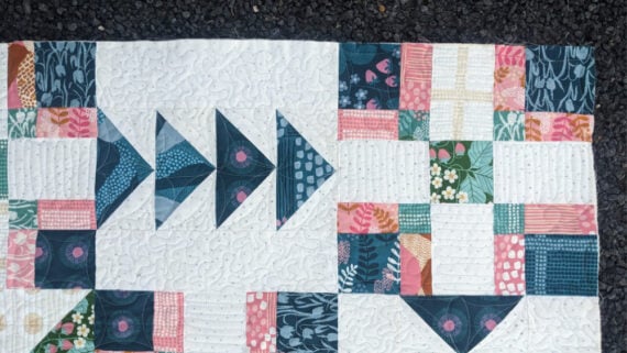 A close-up of a colorful Crosswind Mystery Quilt reveals geometric patterns, featuring blue triangles and patchwork squares in pink, green, and yellow on a white background, lying on a dark textured surface.