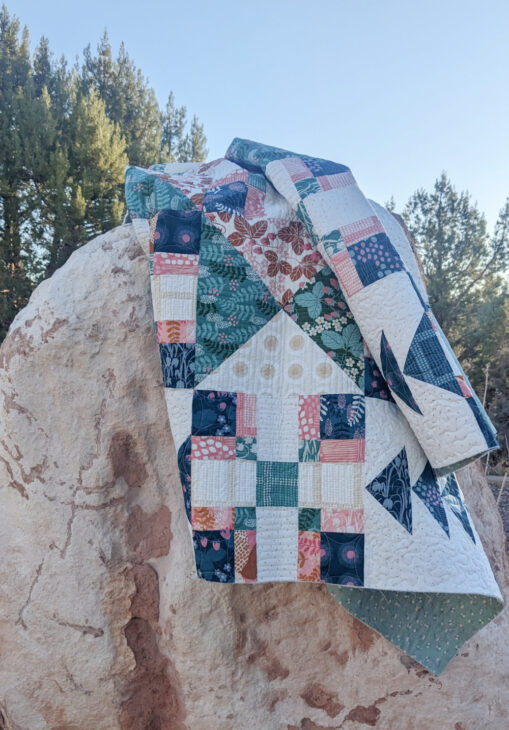 A colorful patchwork quilt, part of the Crosswind Mystery Quilt reveal, with geometric and floral patterns is draped over a large, light-colored rock outdoors, surrounded by pine trees and a clear blue sky.