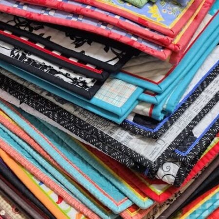A close-up of a stack of colorful, neatly folded quilts and fabric blankets, each featuring different patterns, prints, and bright borders—showcasing edges that wow for a finish it with flair look.