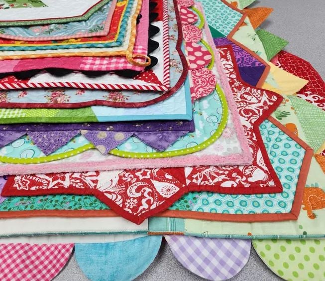 A stack of colorful, patterned quilted table toppers with scalloped edges, neatly layered. Featuring various prints—polka dots, florals, checks—each piece showcases binding basics to finish it with flair.