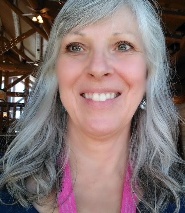 A smiling woman with long, wavy gray hair and light eyes, wearing a pink lanyard and earrings, stands indoors with wooden beams and soft lighting—ready to discuss binding basics and how to finish it with flair.