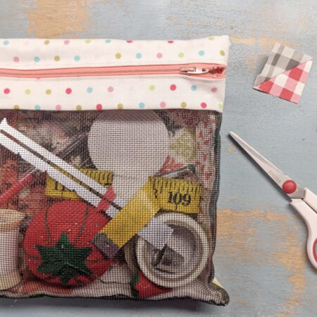 A mesh zipper pouch with a polka-dot top contains sewing supplies like thread, a measuring tape, a pincushion, and scissors. Two patchwork fabric squares from a Quilt Block Tutorial and red-handled scissors are placed beside the pouch.