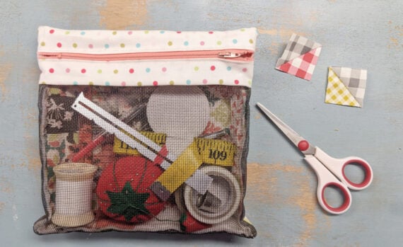 A mesh zipper pouch with a polka-dot top contains sewing supplies like thread, a measuring tape, a pincushion, and scissors. Two patchwork fabric squares from a Quilt Block Tutorial and red-handled scissors are placed beside the pouch.