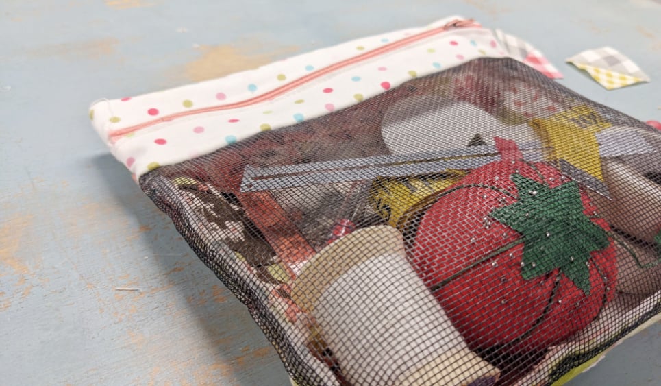 A mesh zippered pouch with a polka dot trim, containing sewing supplies like a red tomato pincushion, thread spools, and an unfinished UFO Quilt Block, sits on a light blue surface.