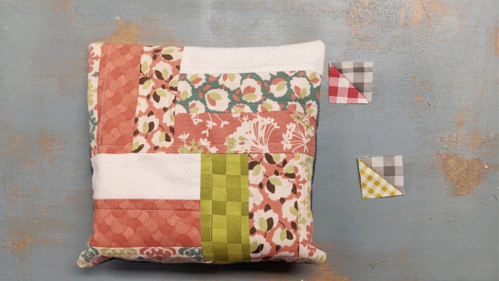 A patchwork pincushion made of various patterned fabrics in pink, green, and white lies on a distressed blue surface&mdash;perfect inspiration for your next Quilt Block Tutorial. Two small plaid triangle fabric pieces rest nearby.