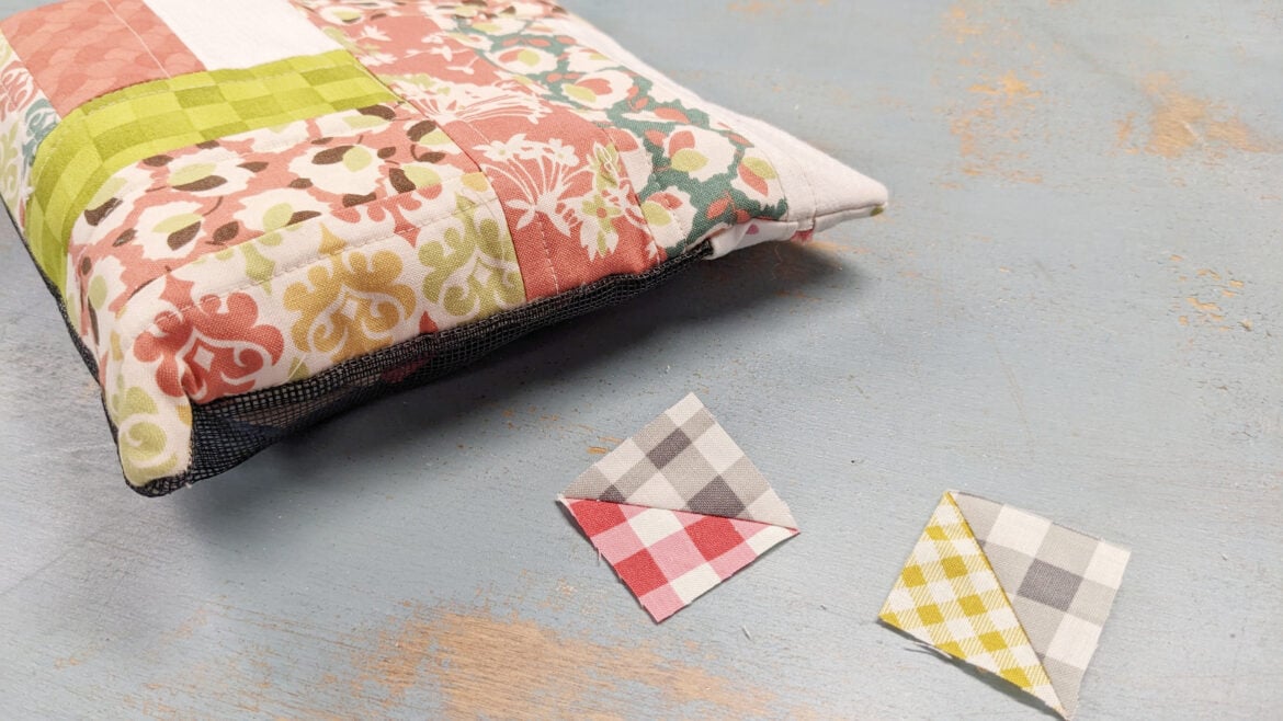 A quilted pouch with floral and checkered fabric patterns sits on a blue surface next to two small patchwork triangles, evoking a UFO Quilt Block design in cheerful pink and yellow checks.