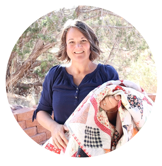 A woman with short brown hair and a navy blue shirt stands outdoors, smiling and holding a colorful patchwork quilt. Trees and a brick structure are visible in the background.