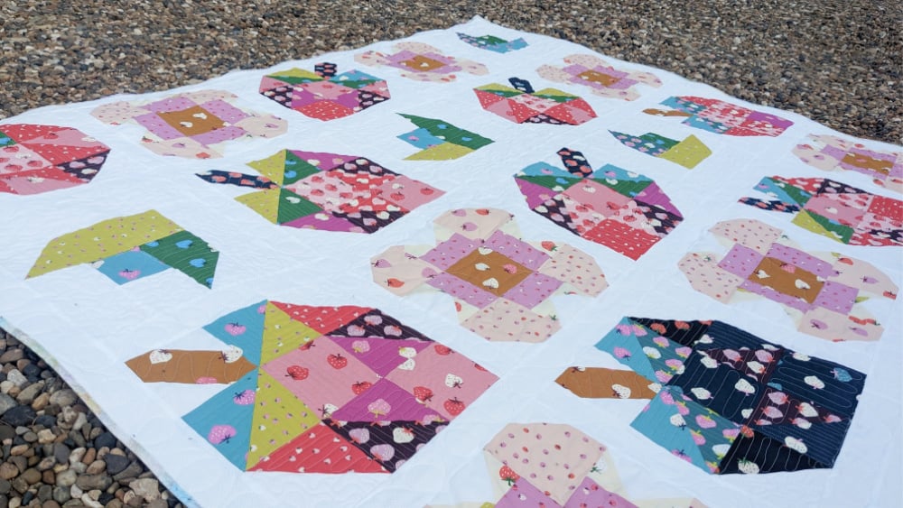 A colorful quilt with a white background is laid on pebbles, featuring various patchwork teapot designs in different patterns and colors&mdash;a charming BOM project to inspire your creativity.