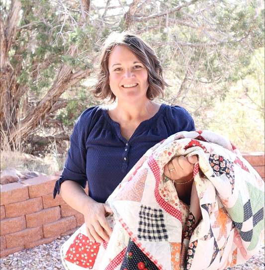 A woman with short brown hair, wearing a blue shirt, smiles while holding a colorful patchwork quilt outdoors in front of trees and a brick wall.
