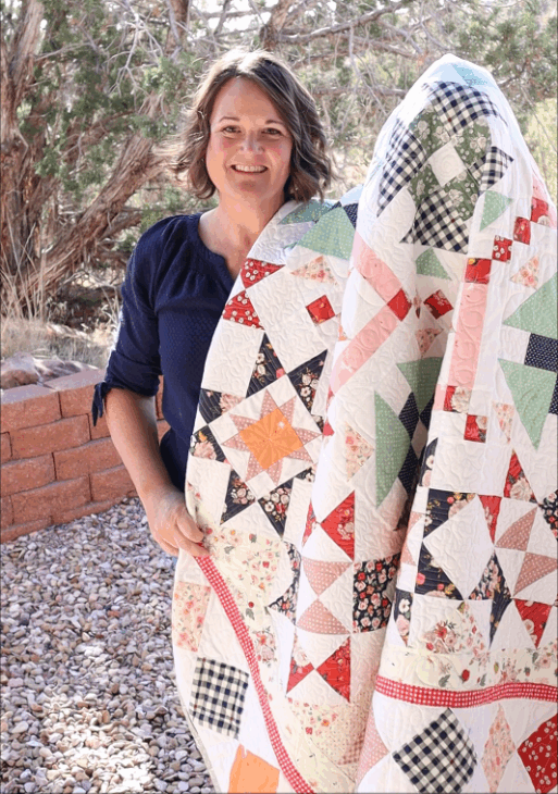 Introducing the Patchworkers Plus 2026 Block of the Month Pattern 8 A woman with short brown hair stands outside holding a colorful patchwork quilt decorated with star patterns from the Starlit Meadow Block of the Month. She is smiling, and trees and a brick wall are visible in the background.