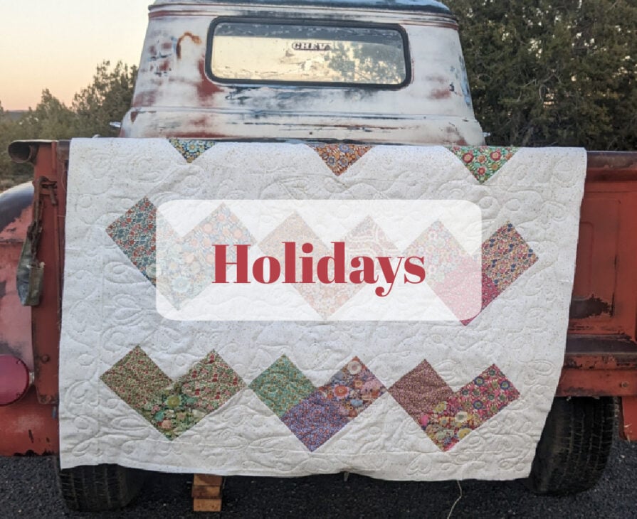 A vintage red truck with a white and patterned quilt draped over its tailgate. The word "Holidays" appears in bold red text over the quilt. Trees are visible in the background.