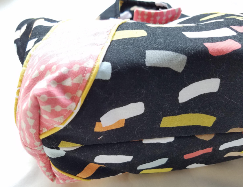 A close-up of a fabric bag using the Iron Caddy Pattern, with a colorful abstract design in white, yellow, grey, and orange on black, plus pink fabric with white dots and yellow piping&mdash;perfect for your next Make & Take Monthly project.