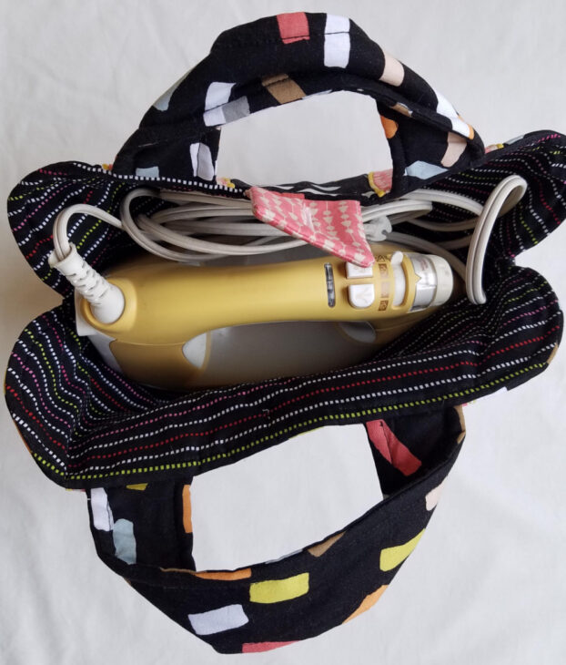 A yellow handheld iron with a white cord is placed inside a black fabric bag made with the Iron Caddy Pattern, featuring colorful rectangles and striped handles, viewed from above. Perfect for your next Make & Take project!.