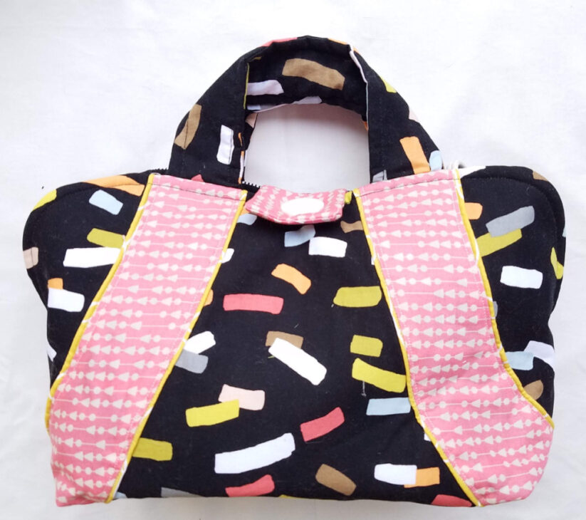 A fabric handbag with multicolored abstract shapes on a black background and wide pink handles featuring white geometric patterns&mdash;perfect for your next Make & Take or as a monthly project using the Iron Caddy Pattern. Displayed on a plain white surface.