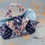 A colorful quilted fabric bag, inspired by the Iron Caddy Pattern, with a plaid handle, blue polka dot lining, and floral prints sits on a distressed blue surface next to scissors and sewing pins—perfect for your next Make & Take project.