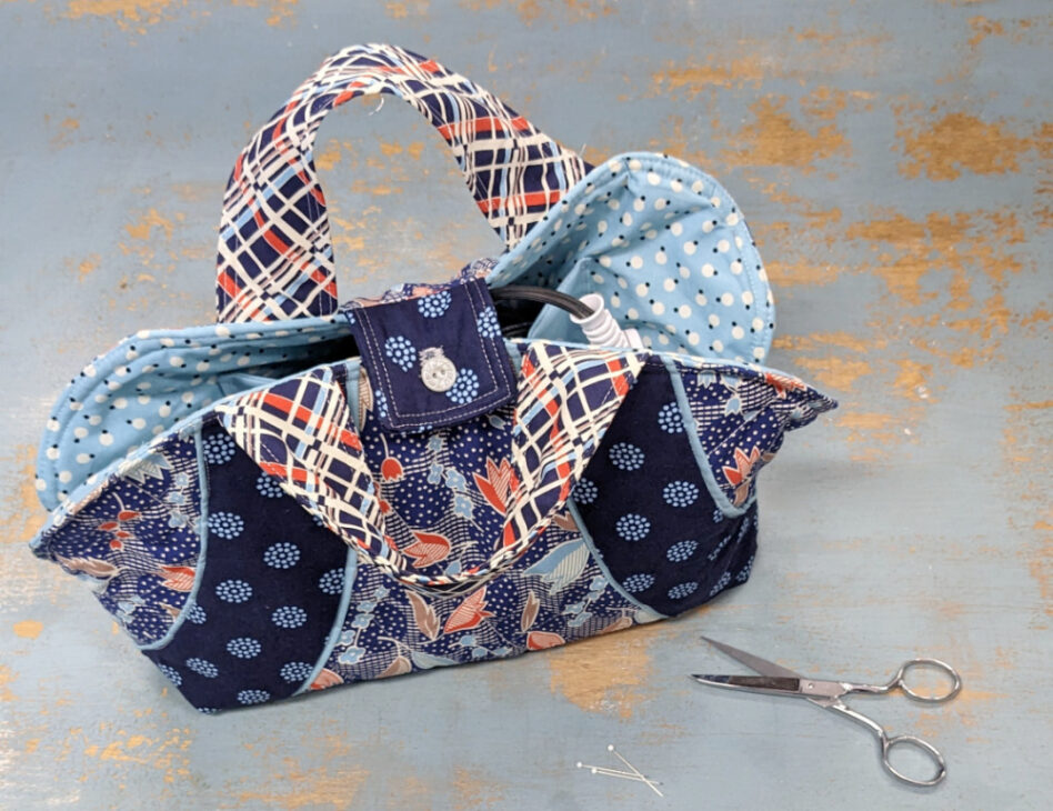 A colorful quilted fabric bag, inspired by the Iron Caddy Pattern, with a plaid handle, blue polka dot lining, and floral prints sits on a distressed blue surface next to scissors and sewing pins&mdash;perfect for your next Make & Take project.