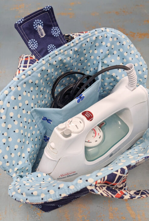 A white iron with a black cord is placed inside a quilted, blue polka-dotted fabric bag made using the Iron Caddy Pattern, featuring a patterned flap and light blue pocket. The background is a distressed wooden surface.