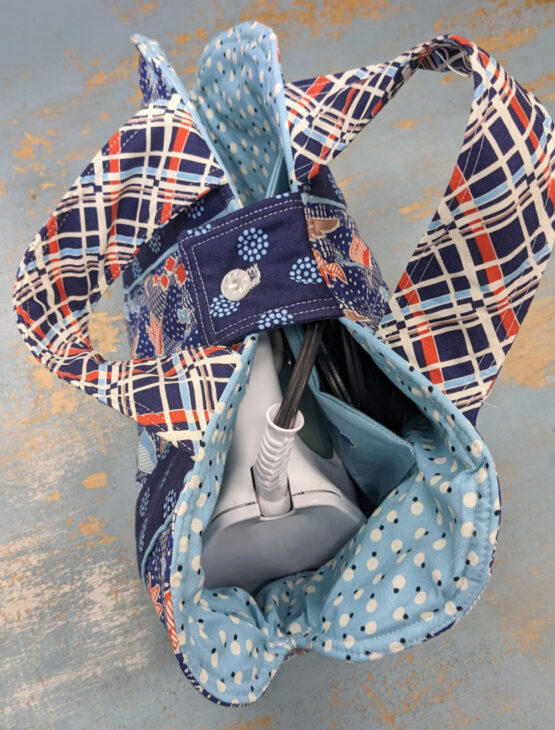 A patterned fabric bag made with the Iron Caddy Pattern, featuring blue, white, and orange designs, opens to reveal a white iron and black cord inside. Perfect for your next Make and Take project on a distressed blue and tan surface.