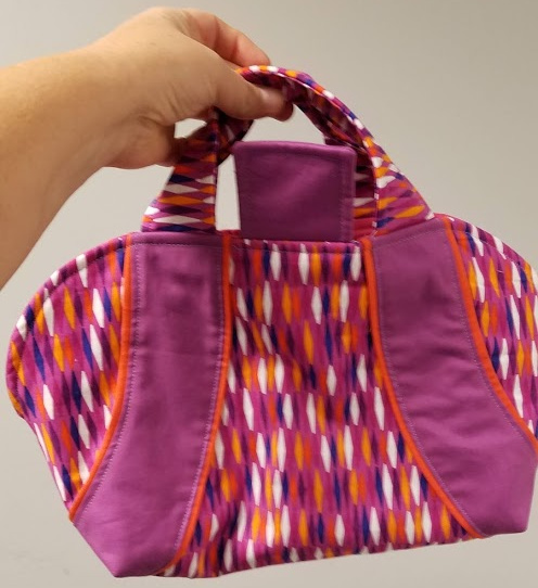 A hand is holding a colorful fabric handbag featuring the Iron Caddy Pattern, with purple sides and a multicolored diamond design in pink, orange, white, and purple on a plain background. Perfect for your next Make & Take session!.
