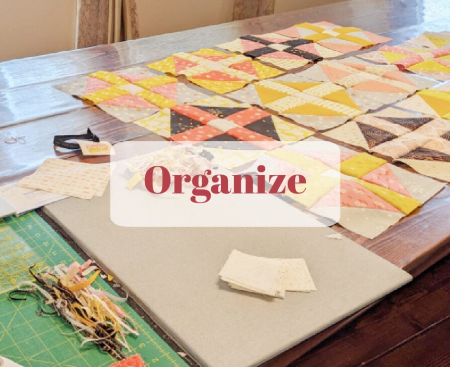 A wooden table covered with colorful fabric quilt squares arranged in rows. A green cutting mat, small fabric scraps, and white cloth squares are nearby. The word "Organize" is overlaid in bold red letters.