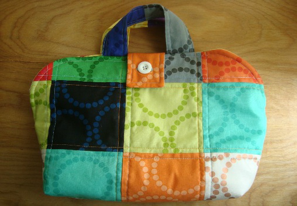 A colorful patchwork fabric bag with short handles, a button closure, and a pattern of dotted circles in bright colors, perfect for a Make & Take or inspired by the Iron Caddy Pattern, placed on a wooden surface.