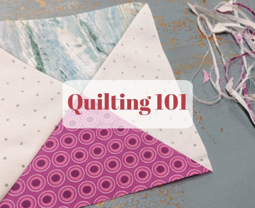 A square quilt block made of white, blue, and pink patterned fabrics sits on a worn blue table with fabric scraps nearby. The words &ldquo;Quilting 101&rdquo; are overlaid in bold red text.