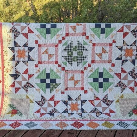 A colorful patchwork quilt with the Starlit Meadow pattern, featuring star and geometric designs in green, orange, blue, yellow, and red fabric draped over a black railing on a wooden deck, with trees in the background. Perfect for any Block of the Month project.