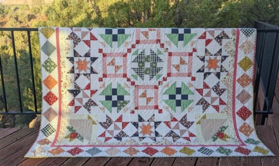 A colorful patchwork quilt with the Starlit Meadow pattern, featuring star and geometric designs in green, orange, blue, yellow, and red fabric draped over a black railing on a wooden deck, with trees in the background. Perfect for any Block of the Month project.