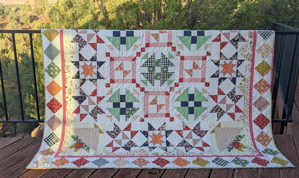 A colorful patchwork quilt with the Starlit Meadow pattern, featuring star and geometric designs in green, orange, blue, yellow, and red fabric draped over a black railing on a wooden deck, with trees in the background. Perfect for any Block of the Month project.