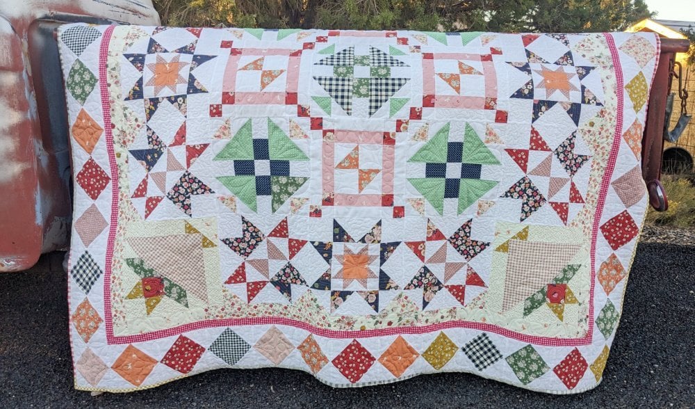 January 2026 BOM + Bonus Iron Caddy 6 A colorful handmade quilt, inspired by the Starlit Meadow Block of the Month 2026, features vibrant star and square patterns in green, orange, pink, and blue fabric, draped over the side of a rustic vehicle outdoors.