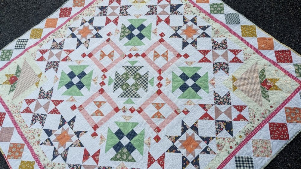 Introducing the Patchworkers Plus 2026 Block of the Month Pattern 7 A colorful patchwork quilt with geometric star and cross patterns, inspired by the Starlit Meadow Block of the Month, in pastel and vibrant fabrics, bordered by a diamond-shaped edge and laid out on a dark surface.