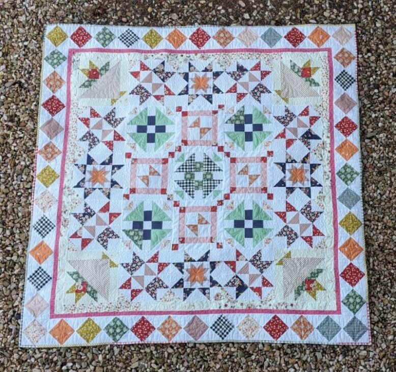 Block of the Month Pattern for 2026 - Starlit Meadow 5 A colorful patchwork quilt, inspired by the Starlit Meadow pattern, features geometric patterns and star shapes in pastel and bright colors. Displayed on gravel, the quilt has multiple borders with intricate square and triangle designs.