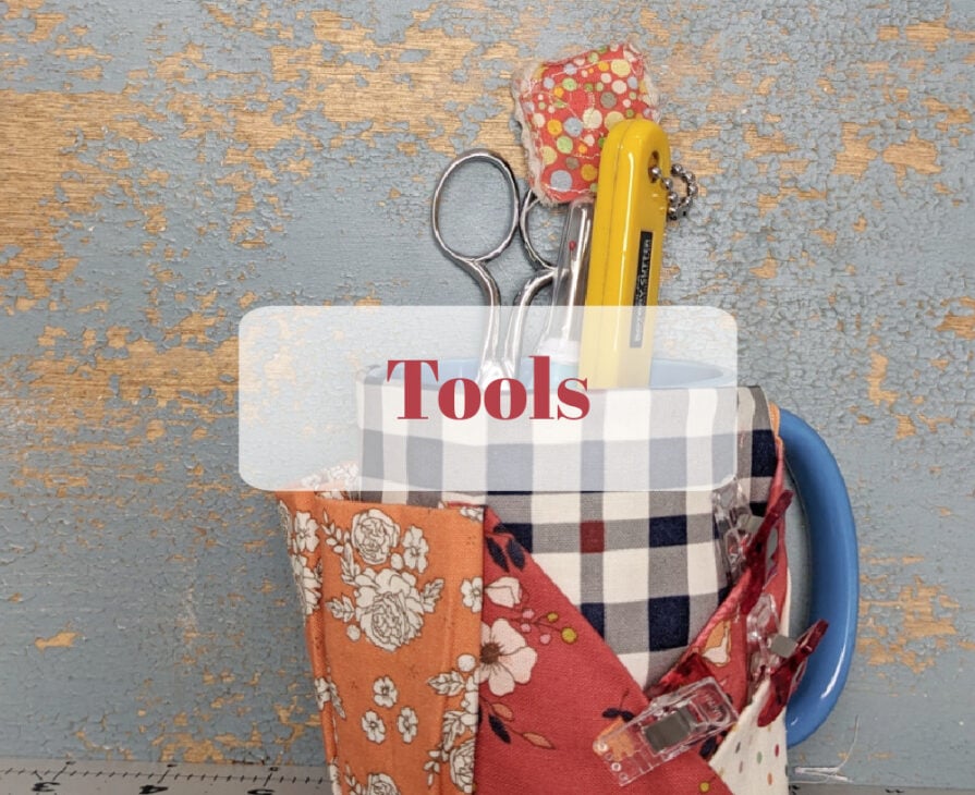 A blue and white mug with a plaid pattern holds sewing tools, including scissors, a rotary cutter, and fabric scraps, against a textured, blue and tan wall. The word "Tools" appears in red letters in the center.