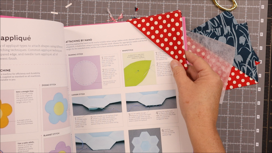 A person holds a red and white polka dot fabric square while reading an open sewing book with illustrated instructions on a cutting mat, working on a 30 minute project. Other fabric pieces for a corner bookmark are scattered nearby.