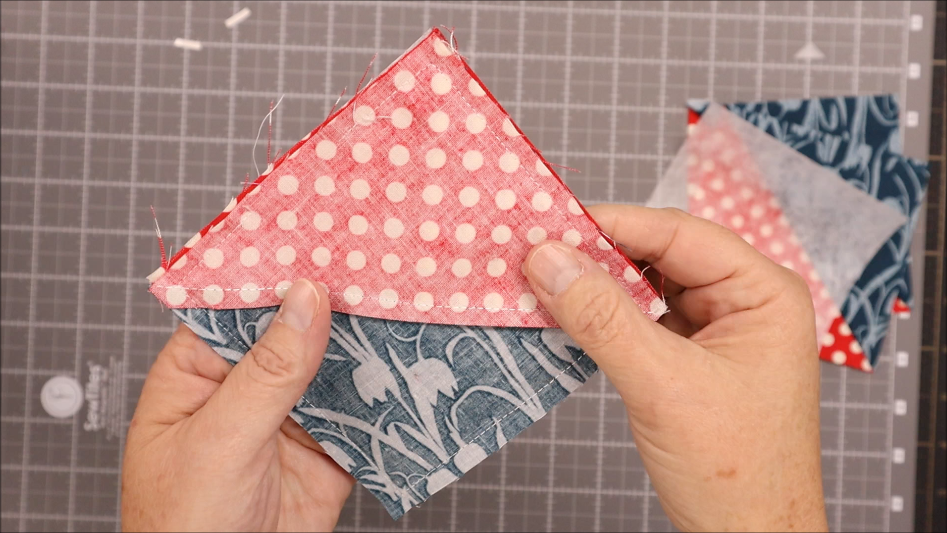 A person holds two sewn fabric pieces&mdash;one pink with white polka dots, the other blue with a floral pattern&mdash;joined along one edge as a corner bookmark, displayed against a grid cutting mat. Perfect for a quick 30 minute project.