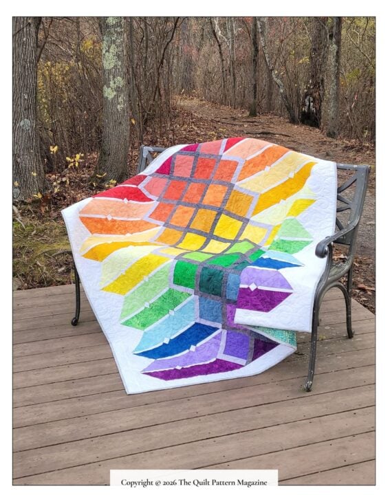 A colorful quilt with a geometric, leaf-like pattern in rainbow colors from a New Issue of Quilt Pattern Magazine is draped over a metal bench on a wooden deck, with bare trees and a forested background.