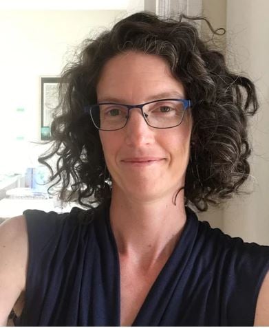 A person with curly brown hair, wearing glasses and a navy blue top, smiles slightly while taking a selfie indoors with light walls and framed artwork&mdash;a cozy scene after attending PW+ Workshops.