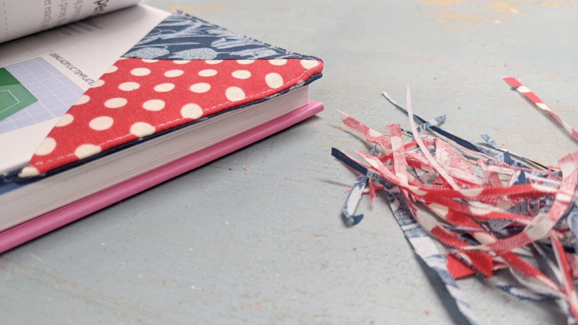 A close-up of a fabric-covered book with a red and white polka dot corner bookmark, lying on a blue surface. Next to it are small piles of multicolored fabric scraps&mdash;perfect inspiration for a quick 30 minute project.