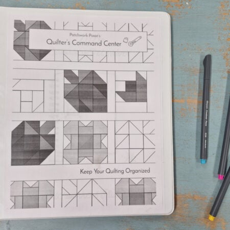 A binder labeled "Quilter's Command Center" with 2026 quilting patterns on the cover sits on a distressed blue surface next to three colored pens: black, blue, and pink.