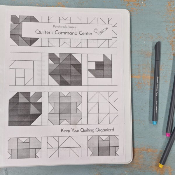 A binder labeled "Quilter's Command Center" with 2026 quilting patterns on the cover sits on a distressed blue surface next to three colored pens: black, blue, and pink.
