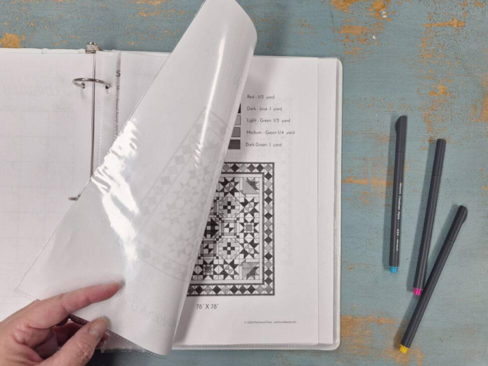 A hand flips a plastic sheet in a Quilter's Command Center binder, revealing a black-and-white quilt pattern and fabric color guide. Three markers&mdash;blue, yellow, and pink&mdash;rest on a distressed blue wooden surface nearby.