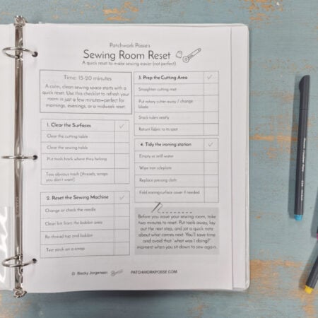An open binder on a blue wooden table displays a printed “Sewing Room Reset” checklist for 2026. Three colored pens (blue, pink, gray) lie nearby as the Quilter's Command Center guides steps for cleaning and organizing your quilting space.