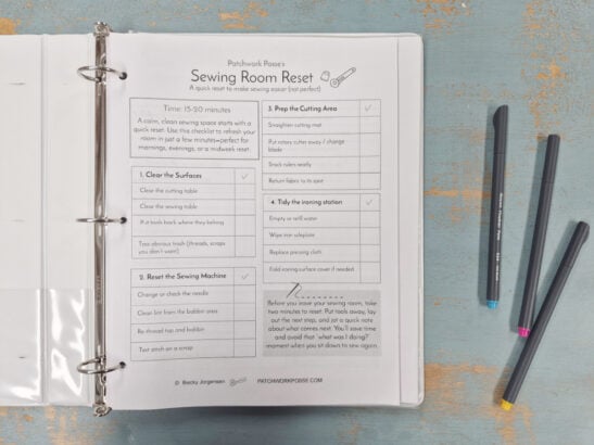 An open binder on a blue wooden table displays a printed &ldquo;Sewing Room Reset&rdquo; checklist for 2026. Three colored pens (blue, pink, gray) lie nearby as the Quilter's Command Center guides steps for cleaning and organizing your quilting space.