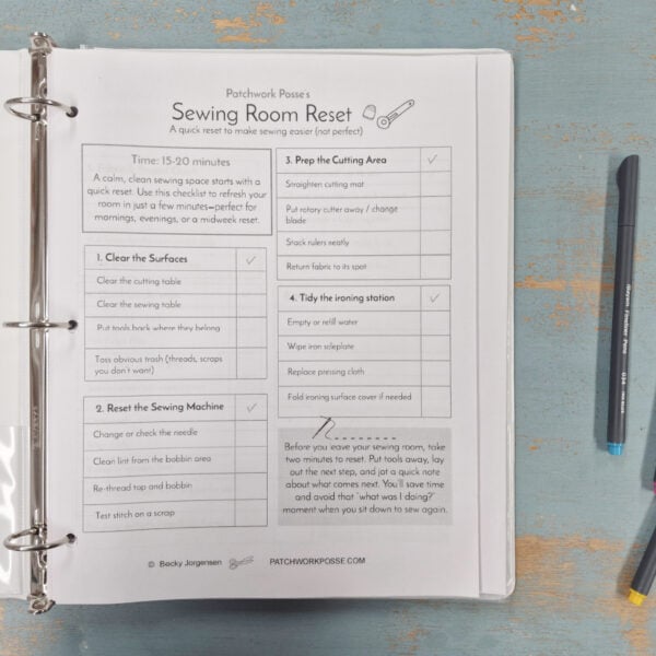 An open binder on a blue wooden table displays a printed “Sewing Room Reset” checklist for 2026. Three colored pens (blue, pink, gray) lie nearby as the Quilter's Command Center guides steps for cleaning and organizing your quilting space.