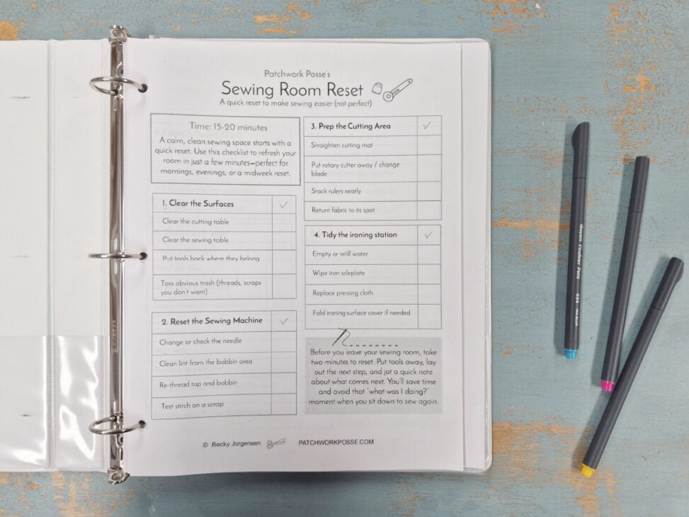 An open binder on a blue wooden table displays a printed &ldquo;Sewing Room Reset&rdquo; checklist for 2026. Three colored pens (blue, pink, gray) lie nearby as the Quilter's Command Center guides steps for cleaning and organizing your quilting space.