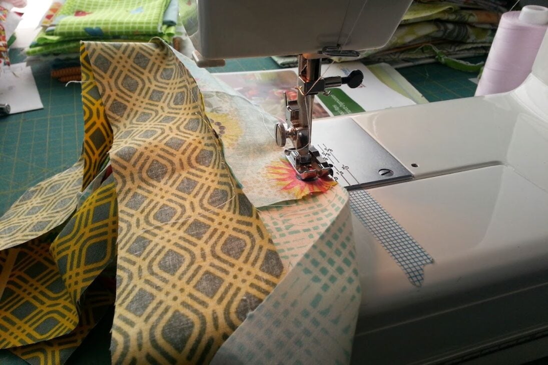Close-up of a sewing machine stitching together colorful patterned fabric strips, with more fabric pieces and sewing supplies in the background.
