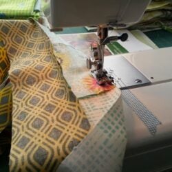Close-up of a sewing machine stitching together colorful patterned fabric strips, with more fabric pieces and sewing supplies in the background.