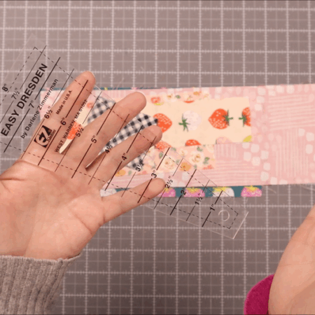 Two hands hold a clear plastic Dresden Plate ruler labeled "Easy Dresden" above a pink, patterned fabric strip on a gray cutting mat with a grid, ready to create beautiful quilt pieces.