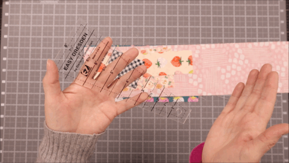 Two hands hold a clear plastic Dresden Plate ruler labeled "Easy Dresden" above a pink, patterned fabric strip on a gray cutting mat with a grid, ready to create beautiful quilt pieces.